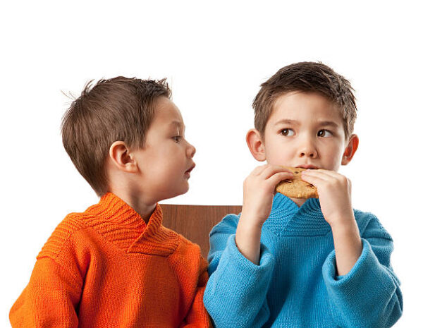 Zwei Jungs im Schulkindalter sitzen nebeneinander und der linke schaut neidisch auf den Keks des rechten Jungen.